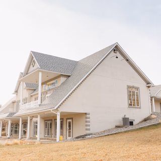 Cream-colored two-story commercial building with a gray shingle roof and white columns, set on a grassy hill.