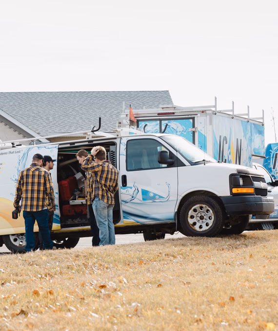 Three service workers in yellow plaid shirts and jeans consult at the open side door of a branded Icon white work van.